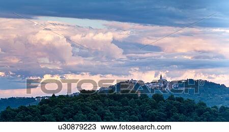 Stock Image - View of Todi hilltop town with church and spire of San Fortunato, at sunset, with bank of trees in foreground, hills beyond, and dramatic billowing grey-blue clouds, panorama view, Umbria, Italy. Fotosearch