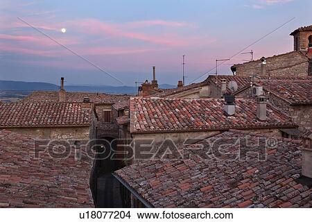 Picture - View over traditionally red-tiled terracotta rooftops and narrow street of medieval stone village to hills beyond Tiber valley at sunset, with rising full moon in pink and blue sky, Umbria, Italy. Fotosearch