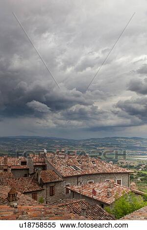 Stock Photography - View over traditionally red-tiled terracotta rooftops of medieval stone village to fields of Tiber valley and hills beyond, with threatening billowing grey clouds, Umbria, Italy. Fotosearch