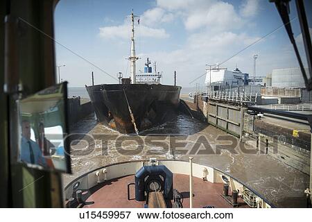 Stock Photo - Tugboat pulling ship in urban harbor. Fotosearch