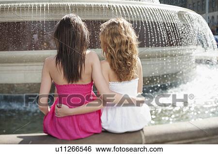 Back view of two young female friends with arms around sitting by water fountain View Large Photo Image Stock Photograph - Back view of two young female friends with arms around sitting by water fountain. Fotosearch