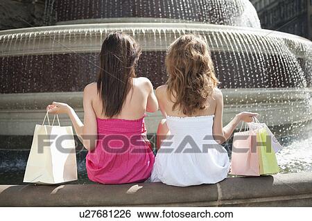 Back view of two young female friends with shopping bags sitting by water fountain View Large Photo Image Stock Photograph - Back view of two young female friends with shopping bags sitting by water fountain. Fotosearch