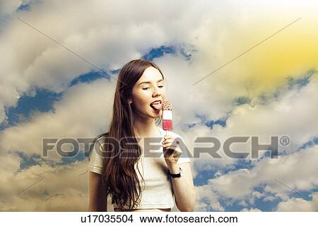 Beautiful young woman licking popsicle against cloudy sky View Large Photo Image Picture - Beautiful young woman licking popsicle against cloudy sky. Fotosearch