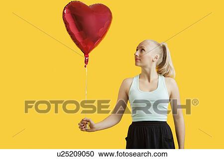Beautiful young woman looking at heart shaped balloon over yellow background View Large Photo Image Stock Image - Beautiful young woman looking at heart shaped balloon over yellow background. Fotosearch