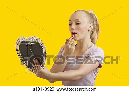 Beautiful young woman looking at mirror while applying lipstick over yellow background View Large Photo Image Stock Photo - Beautiful young woman looking at mirror while applying lipstick over yellow background. Fotosearch