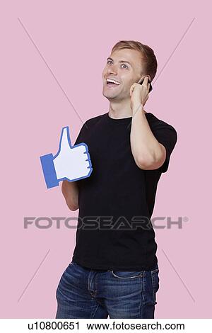 Stock Image - Cheerful young man using cell phone while holding fake like button over pink background. Fotosearch