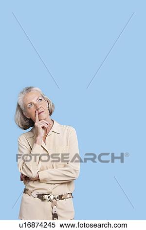 Stock Photography - Contemplative senior woman in casuals with hand on chin against blue background. Fotosearch