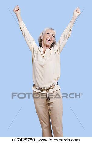 Stock Image - Excited senior woman in casuals cheering with arms raised against blue background. Fotosearch