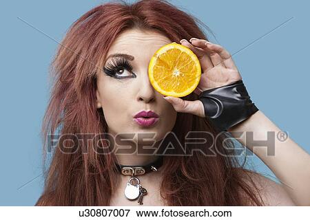 Stock Photo - Funky young woman covering eye with sliced orange over blue background. Fotosearch
