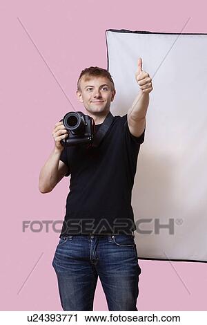 Stock Image - Happy photographer holding camera with thumbs up gesture over pink background. Fotosearch