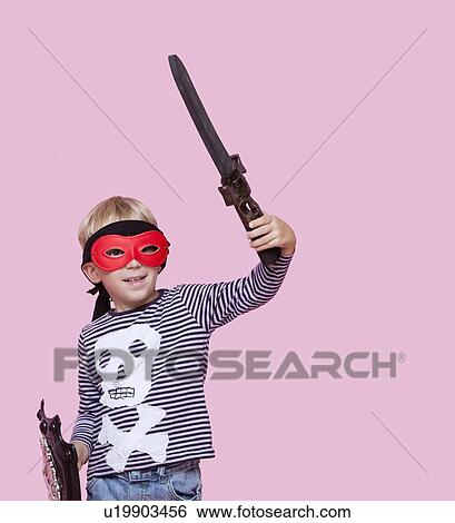 Stock Photograph - Happy young boy wearing eye mask holding sword and shield over pink background. Fotosearch