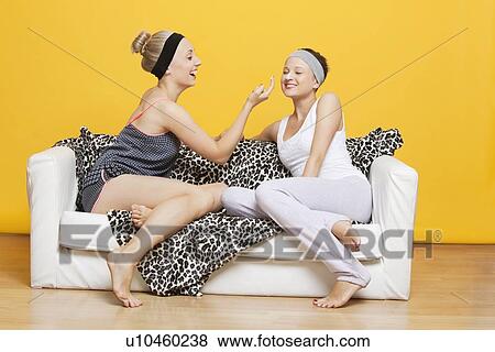 Happy young woman applying face pack on friend's face while sitting on sofa against yellow wall View Large Photo Image Stock Photo - Happy young woman applying face pack on friend's face while sitting on sofa against yellow wall. Fotosearch