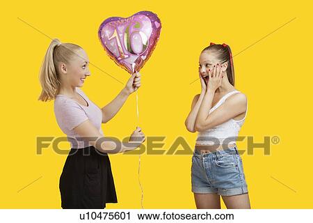 Stock Image - Happy young woman giving birthday balloon to amazed woman standing over yellow background. Fotosearch