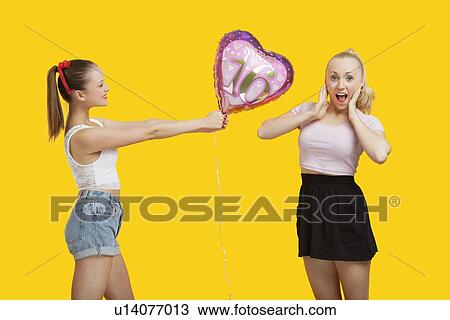 Happy young woman giving birthday balloon to amazed woman standing over yellow background View Large Photo Image Stock Image - Happy young woman giving birthday balloon to amazed woman standing over yellow background. Fotosearch