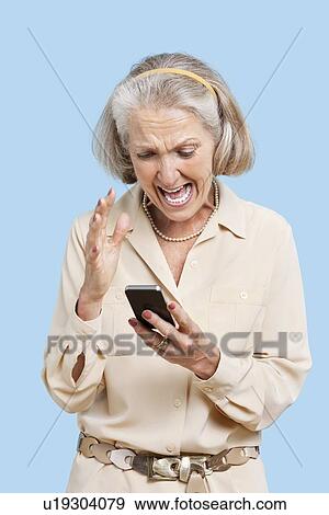 Stock Photo - Irritated senior woman reading text message on cell phone against blue background. Fotosearch