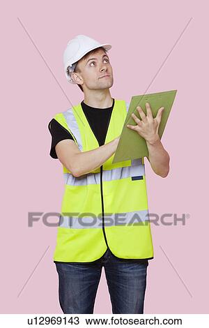 Stock Image - Male construction worker with clipboard looking away over pink background. Fotosearch
