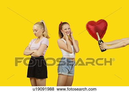 Stock Photo - Man's hand gifting heart shaped balloon and champagne bottle to surprised woman with friend feeling left out standing behind. Fotosearch
