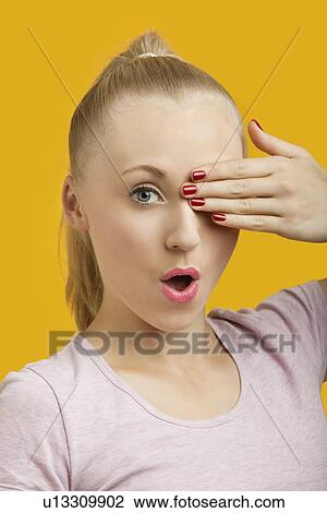 Stock Image - Portrait of a beautiful young woman covering eye over yellow background. Fotosearch