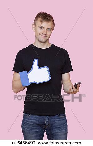 Portrait of a happy young man with cell phone holding fake like button over pink background View Large Photo Image Stock Photo - Portrait of a happy young man with cell phone holding fake like button over pink background. Fotosearch