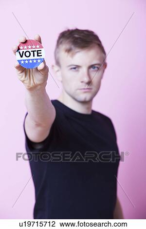 Portrait of a young man holding a VOTE badge up to the camera against pink background View Large Photo Image Stock Image - Portrait of a young man holding a VOTE badge up to the camera against pink background. Fotosearch