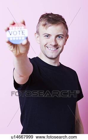 Portrait of a young man holding a VOTE badge up to the camera against pink background View Large Photo Image Stock Photo - Portrait of a young man holding a VOTE badge up to the camera against pink background. Fotosearch