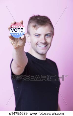 Portrait of a young man holding a VOTE badge up to the camera against pink background View Large Photo Image Stock Photo - Portrait of a young man holding a VOTE badge up to the camera against pink background. Fotosearch