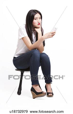 Stock Photo - Portrait of beautiful young woman in casuals sitting on stool with mobile phone against white background. Fotosearch