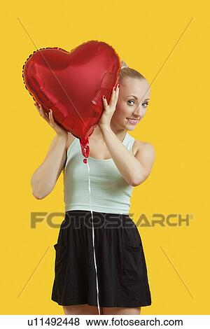Portrait of beautiful young woman peeking through heart shaped balloon over yellow background View Large Photo Image Stock Photo - Portrait of beautiful young woman peeking through heart shaped balloon over yellow background. Fotosearch