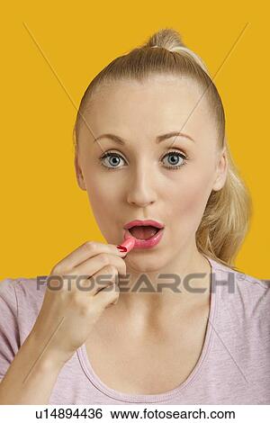 Stock Photograph - Portrait of beautiful young woman applying lipstick over yellow background. Fotosearch