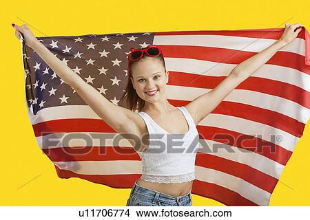 Picture - Portrait of happy young woman holding American flag over yellow background. Fotosearch