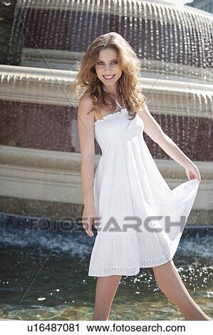 Stock Image - Portrait of happy young woman in white halter neck dress standing by water fountain. Fotosearch