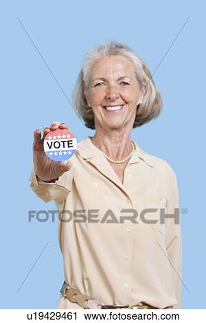 Stock Image - Portrait of senior woman holding an election badge against blue background. Fotosearch