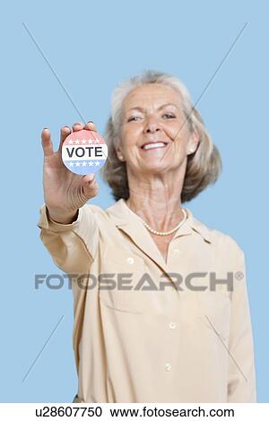 Stock Image - Portrait of senior woman holding an election badge against blue background. Fotosearch