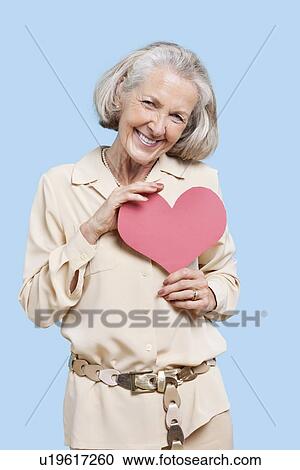 Portrait of senior woman holding red paper heart against blue background View Large Photo Image Stock Image - Portrait of senior woman holding red paper heart against blue background. Fotosearch