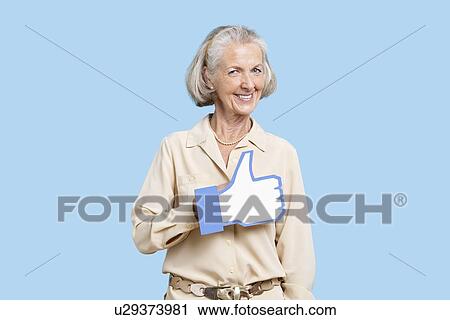 Stock Image - Portrait of senior woman in casuals holding fake like button against blue background. Fotosearch