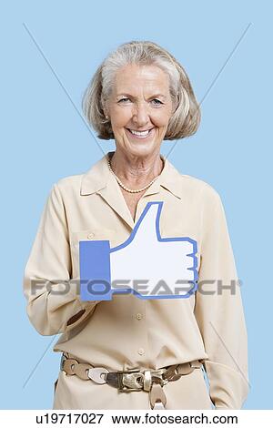 Stock Photo - Portrait of senior woman in casuals holding fake like button against blue background. Fotosearch