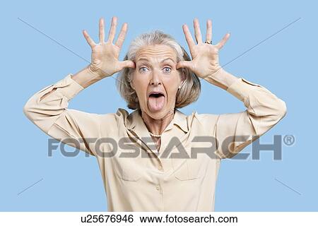 Portrait of senior woman making funny faces with hands on head against blue background View Large Photo Image Stock Photograph - Portrait of senior woman making funny faces with hands on head against blue background. Fotosearch