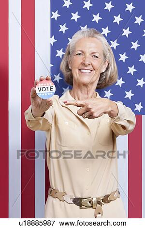 Stock Photo - Portrait of senior woman pointing at election badge against American flag. Fotosearch