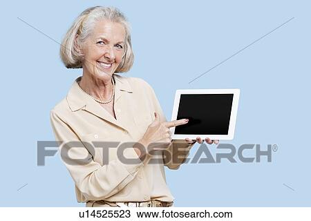 Stock Image - Portrait of senior woman showing tablet PC against blue background. Fotosearch