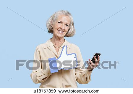 Stock Photo - Portrait of senior woman with cell phone holding fake like button against blue background. Fotosearch