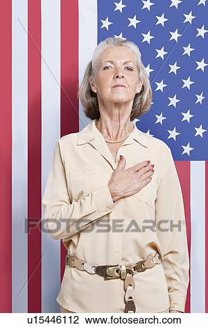 Stock Image - Portrait of senior woman with hand over heart against American flag. Fotosearch