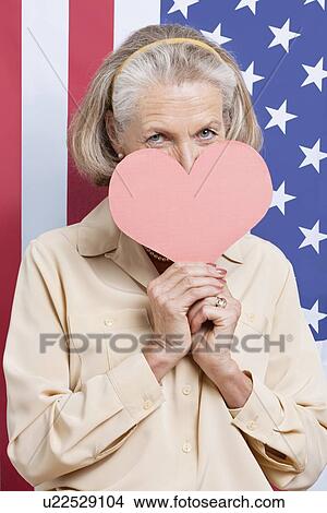 Picture - Portrait of senior woman with red paper heart against American flag. Fotosearch