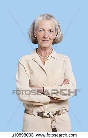 Stock Image - Portrait of smiling senior woman in casuals with arms crossed against blue background. Fotosearch