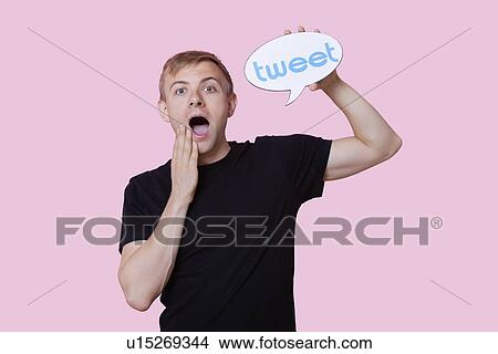 Picture - Portrait of surprised young man holding tweet bubble against pink background. Fotosearch