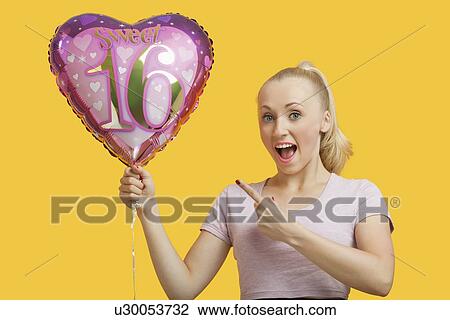 Stock Image - Portrait of surprised young woman holding heart shaped birthday balloon over yellow background. Fotosearch