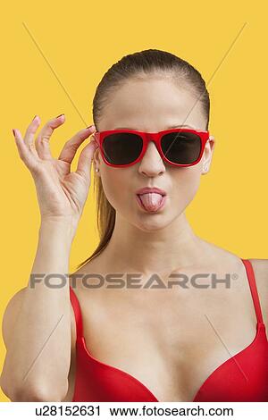 Stock Image - Portrait of young woman in sunglasses sticking out tongue over yellow background. Fotosearch
