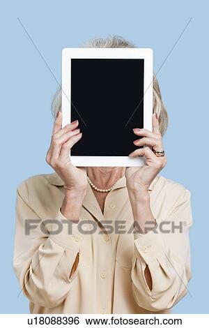 Stock Photograph - Senior woman holding tablet PC in front of her face against blue background. Fotosearch