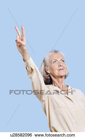 Stock Photograph - Senior woman in casuals gesturing peace sign against blue background. Fotosearch