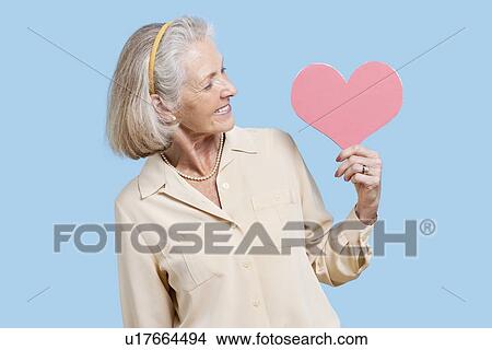 Senior woman in casuals holding red paper heart against blue background View Large Photo Image Picture - Senior woman in casuals holding red paper heart against blue background. Fotosearch