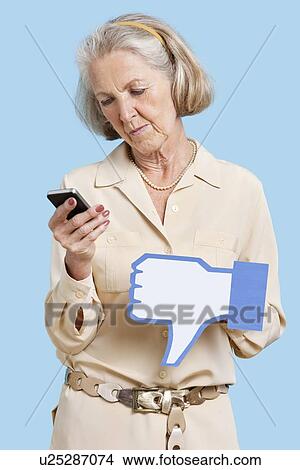 Picture - Senior woman with cell phone holding fake dislike button against blue background. Fotosearch
